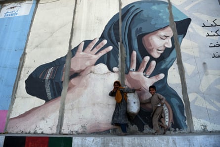 Children carrying a sack of garbage walk past a mural portraying an anguished woman on a roadside in Kabul