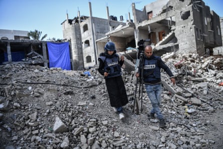 Reba Khalid al-Ajami (L) walks across rubble alongside a cameraman who is carrying his camera and tripod over his shoulder. Both are wearing ‘press’ flak jackets, Ajami also has a helmet