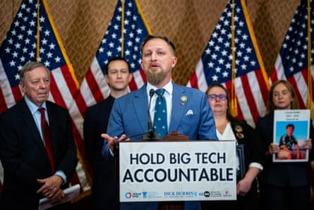 A man in a blue suit and a blue tie holding a sign saying ‘Hold Big Tech Accountable’ with a row of people and stars and stripes flags behind him