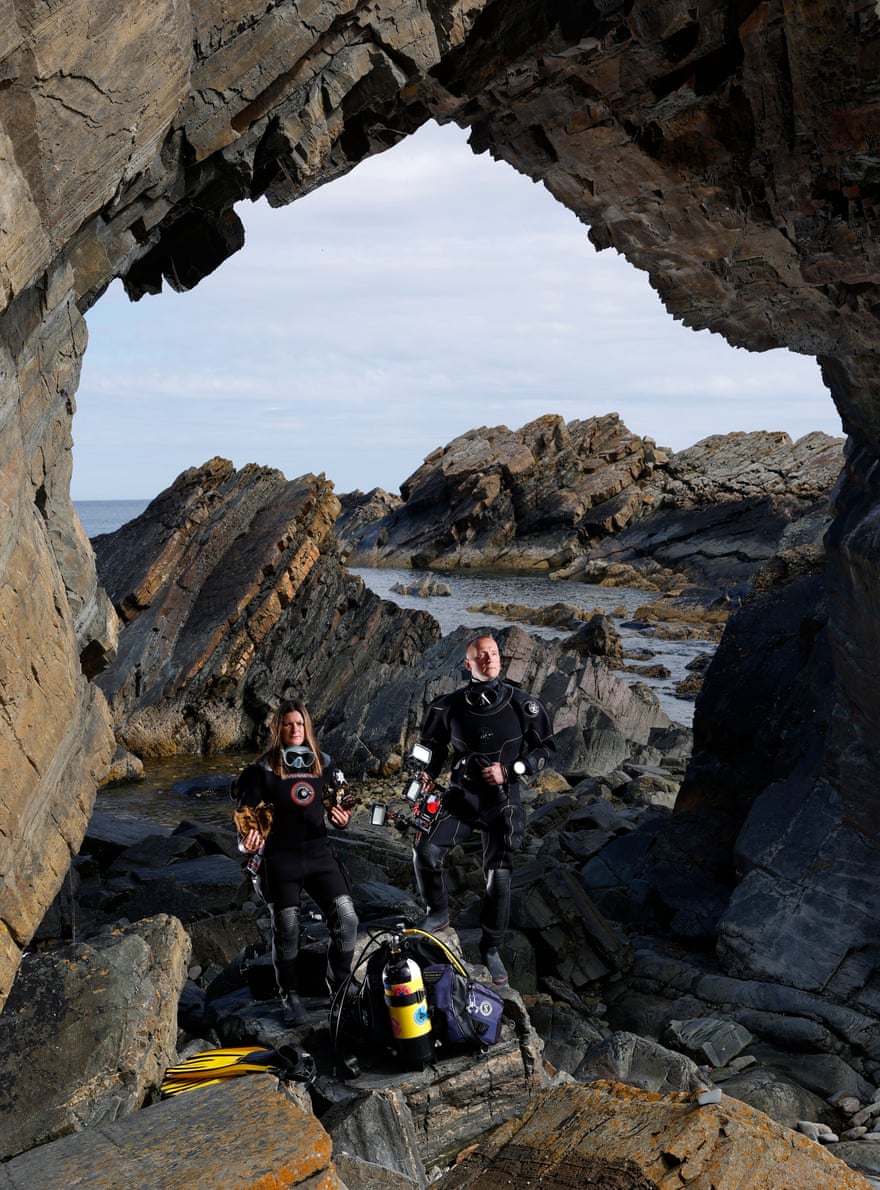 A woman and a man in scuba-diving drysuits pose in a stone arch by a bay 