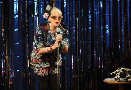 A woman in a colourful, sparkly jacket and sunglasses stands at a mic on stage.