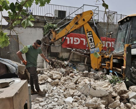 A man in a green T shirt stands amid rubble of a destroyed house by a yellow JCB digger.
