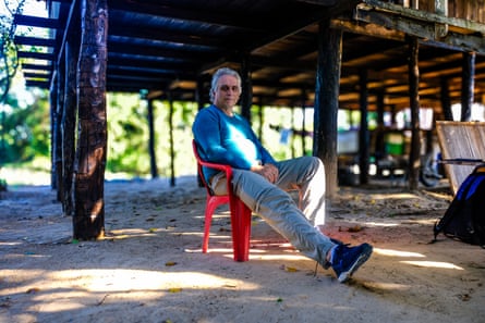 Um homem branco mais velho está sentado em uma cadeira sob um edifício sobre palafitas.
