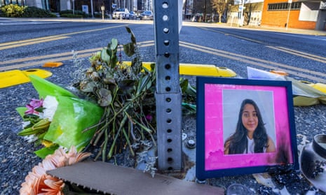 A closeup shot of a memorial at the bottom of a signpost in the middle of an intersection, with a framed photo with a bright pink mat of a smiling young Indian woman with long black hair, a candle, and dried bouquets of flowers.