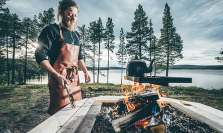 A young bearded man in an apron cooking outdoors over a fire, a lake and trees in the background