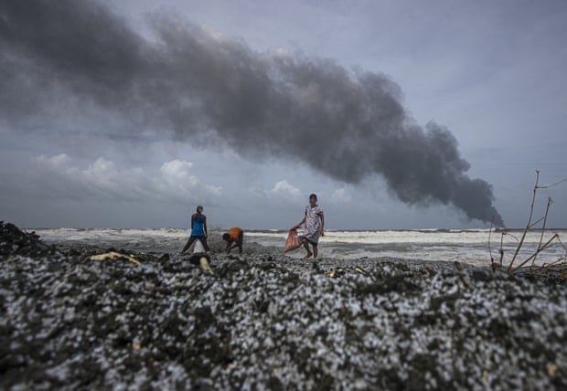 Sri Lankans walk along a beach covered in plastic pellets with smoke rising from a ship wreck in the distance