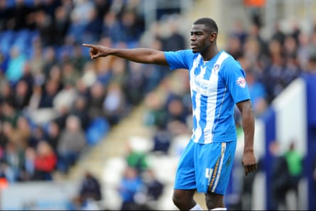 Magnus Okuonghae playing for Colchester United; he wears a blue and white striped top and blue shorts, and is gesticulating with one hand held out.