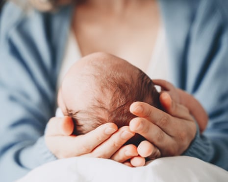 A mother's hands cradle the head of her newborn baby. The baby has thin, dark hair. The mother wears a pale blue gown.