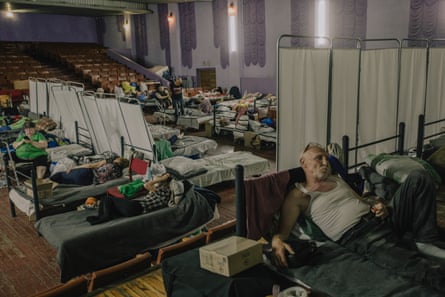 People sleeping on makeshift cots in a former concert hall with a wooden floor