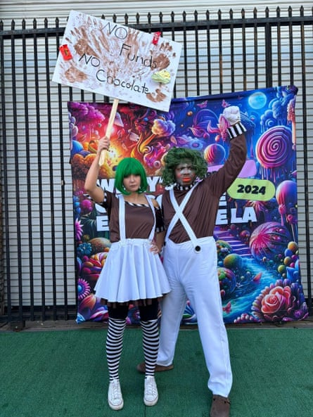 woman and man in green wigs stand in front of tapestry saying ‘willy’s chocolate experience LA’. the woman is holding a sign saying ‘no funds no chocolate’ - it’s covered in chocolate