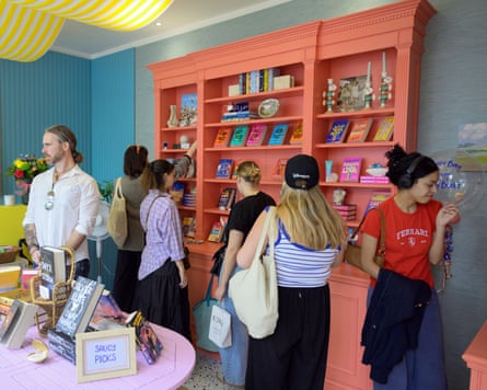 A group of mostly young women browse books displayed on a coral bookcase. The shop is decorated is pastel colours