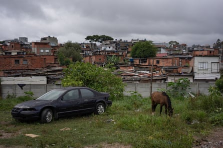 A horse grazes near an abandoned car at the edge of a group of makeshift houses.