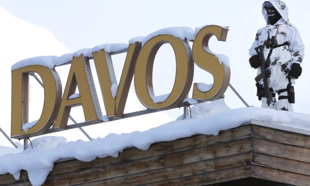 An armed officer is snow camouflage stands guard on a snow-covered roof by the big Davos sign