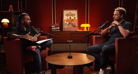 Rajan and Southgate sit opposite each other in soft chairs, across a low table, in a warm, cosy red room with dim lighting