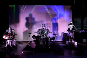 three humanoid robots are lined up on a stage, two with guitars and one sitting behind a drum kit