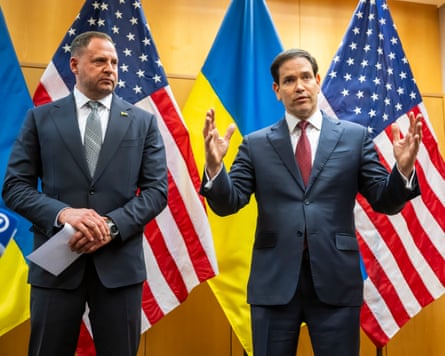 Andriy Yermak stands next to Marco Rubio as Rubio speaks with his hands outstretched; there are furled US and Ukrainian flags behind them. Both men wear suits and ties.