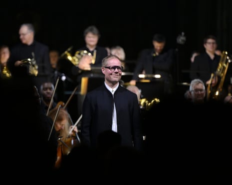 In glasses, a white shirt and black jacket, Max Richter stands onstage while behind him, in softer focus, sit the musicians who have performed his score for Hamnet
