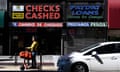 On a sunny urban street, a person rides a scooter past two storefronts, one with a red sign that says 'Checks Cashed' and the other with a blue sign that says 'Payday Loans'.