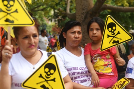 A woman holds a child who holds a triangular yellow placard with a skull and crossbones partly made from the words ‘Mineria metalica’. Another female protester holds the same sign in front.