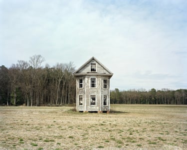 A neat clapboard house, weathered grey, with bay windows and a loft room under its pitched roof, sits in a large scrubby field. Dead creepers cling to the exterior