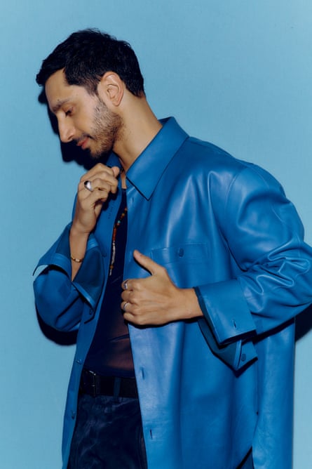 Riz Ahmed photographed from the side in a blue leather shirt with dark top and trousers, while looking down.