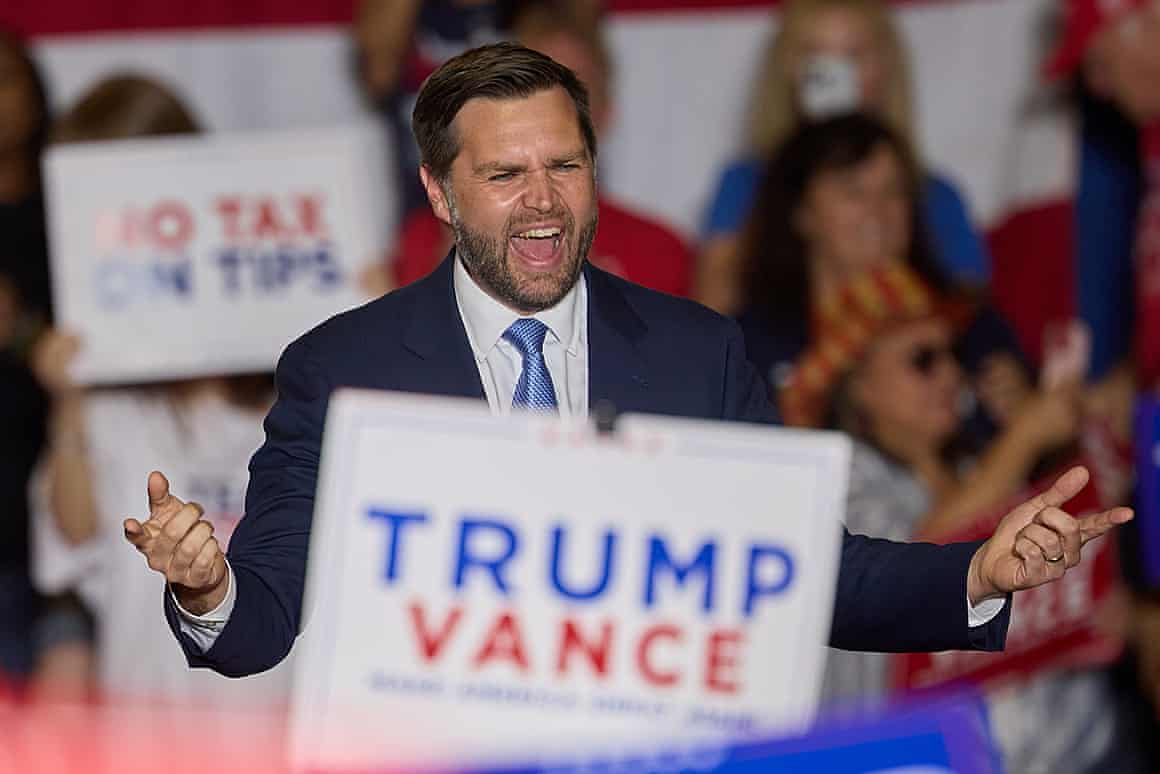 Vance gestures behind a ‘Trump/Vance’ placard