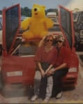 A man with sunglasses, a red shirt and jeans sits on the hood of a sports car with a woman in a matching outfit on his lap. There is a large stuffed bear on the car’s roof, and the vehicle’s butterfly-style doors are open.