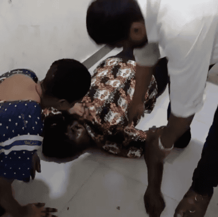 Two people stand over a woman lying on the floor
