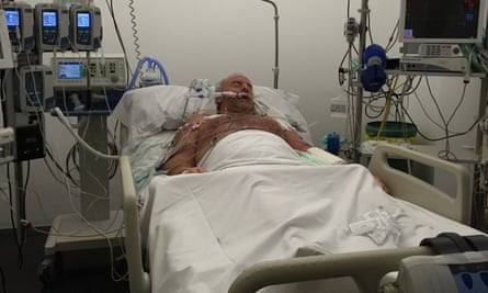 A mid-distance photo of a man lying in a hospital bed connected to numerous medical devices