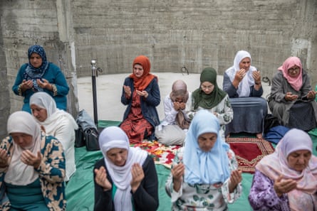 Eleven women, wearing headscarves of various colours, kneel in prayer.