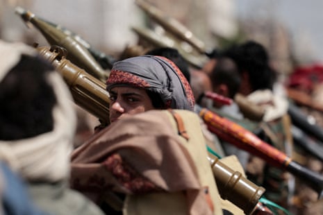 A person wearing a patterned headscarf holds a rifle among other armed individuals