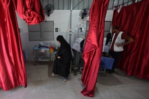 A woman sits on a hospital bed while medical staff attend to a patient in the bed next to her
