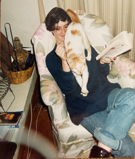Young Emma sits in an armchair, trying to read a book while a cat crawls all over her