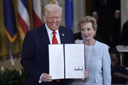 A man wearing a red tie smiles and holds up a document next to a woman in a blue pantsuit