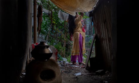 A middle-aged woman in a shalwar kameez [trousers and tunic] standing outside a corrugated iron-walled shack. A clay oven is in the gloomy interior