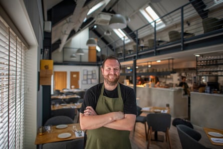 he has a gingery beard and wears a black top with khaki apron. The restaurant is in a barn-like building and is modern-looking with a high arched roof and upper layer of tables on a balcony over the bar and kitchen area