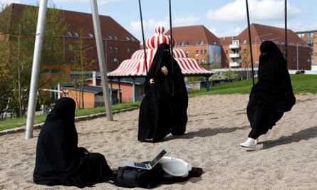 Two women on swing and one sitting in sand of play park, all dressed in black burqas