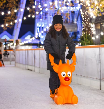 A child skating at night with a mini-reindeer skate aid, with Christmas lights in the background