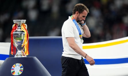 Gareth Southgate, bearded and wearing a T-shirt and a medal on a thick ribbon that reads “UEFA EURO 2024”, walks past a trophy and looks sadly at the ground