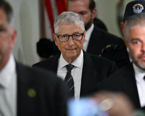 Bill Gates with grey hair and black-framed glasses in a dark suit looks toward the camera