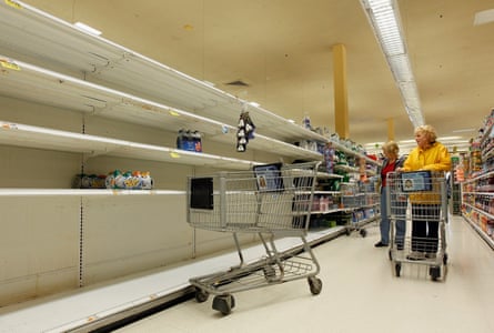 A woman pushing a shopping trolley grabs the last water bottles from a long empty shelf in a supermarket.