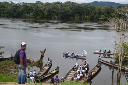 A man in a cap stands on a riverbank while people sit in canoes on the water below