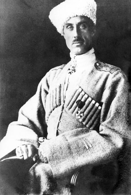 Black-and-white photo of a white man with a long face and a mustache and light-colored eyes, looking directly at the camera, posed and sitting tall with a high white perhaps fuzzy hats, a turtleneck with an Iron Cross on a necklace, and a large, baggy-sleeved military coat.