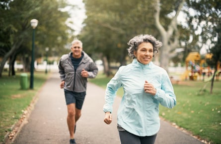 A mature man and woman wearing waterproof running gear smile as they run through a park