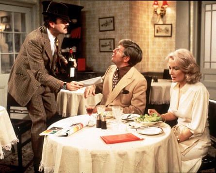 Cleese stands in hat next to couple at dining table