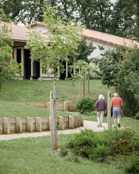 two older people walking along a curving path  past newly planted trees towards a building with a clay-tiled roof
