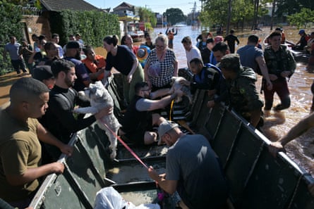 People wade down a flooded street towards a boat. People are getting into the boat and others are passing pet dogs into it.