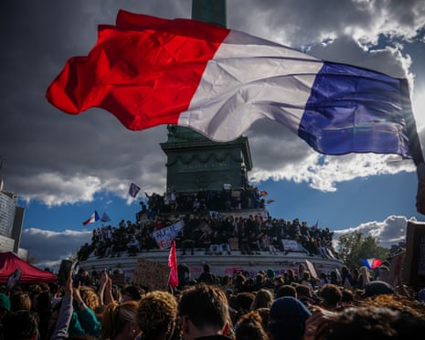 A large French flag waves over protesters gathered around a monument during a school protest