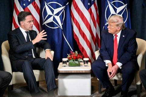 Two men sitting in matching armchairs on a stage speaking in front of two US flags and two Nato flags (a white compass on a blue background).