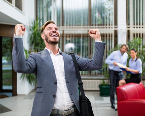 Young businessman celebrating success with arms raised. There is a blurred image of two coworkers in the background.